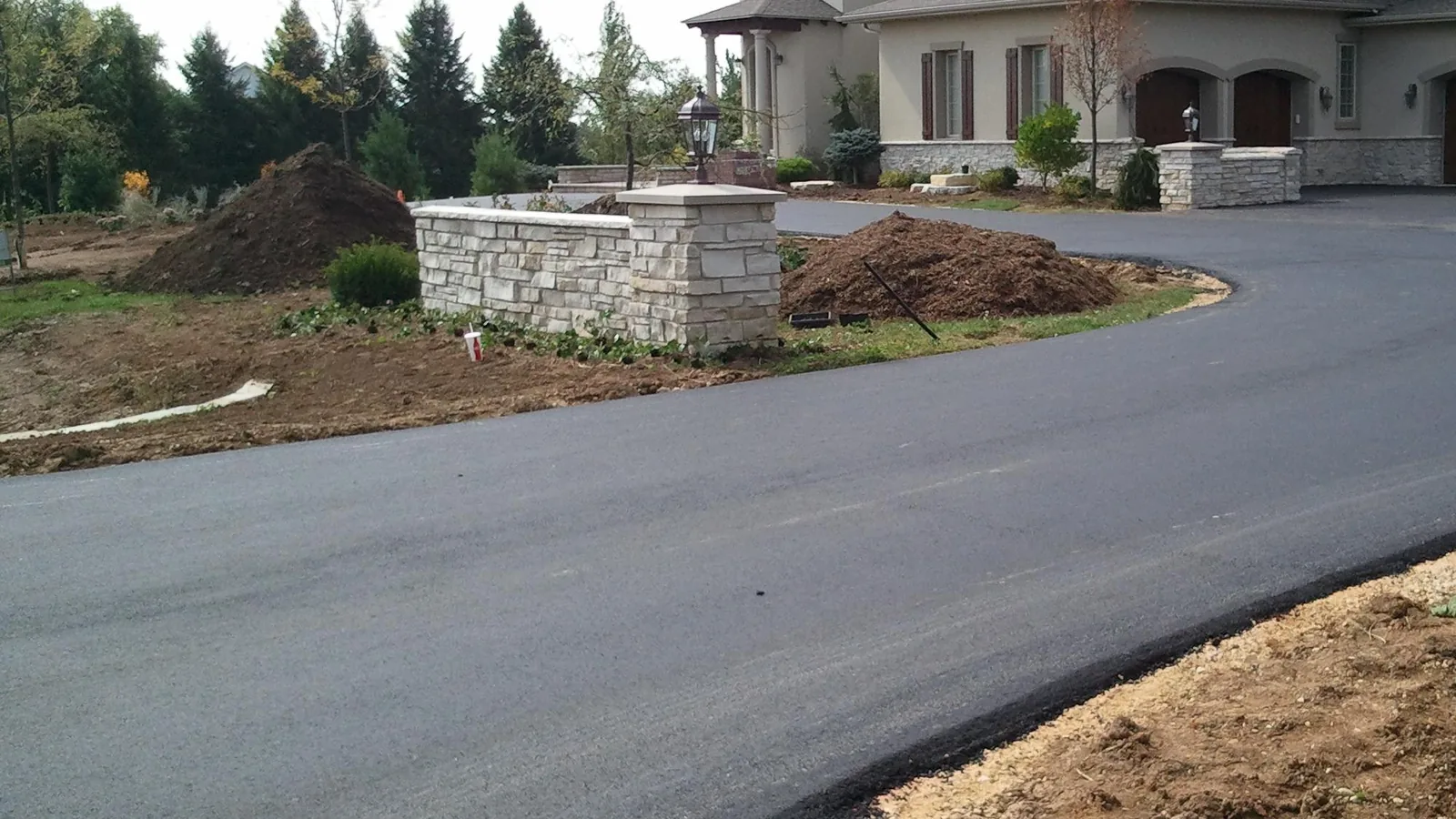Wide new asphalt driveway curving past stone columns to a large home